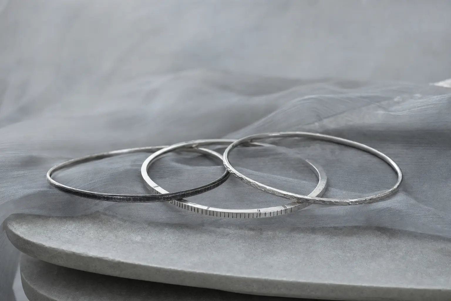 Slim silver bangles loosely overlapping on a rounded gray ledge