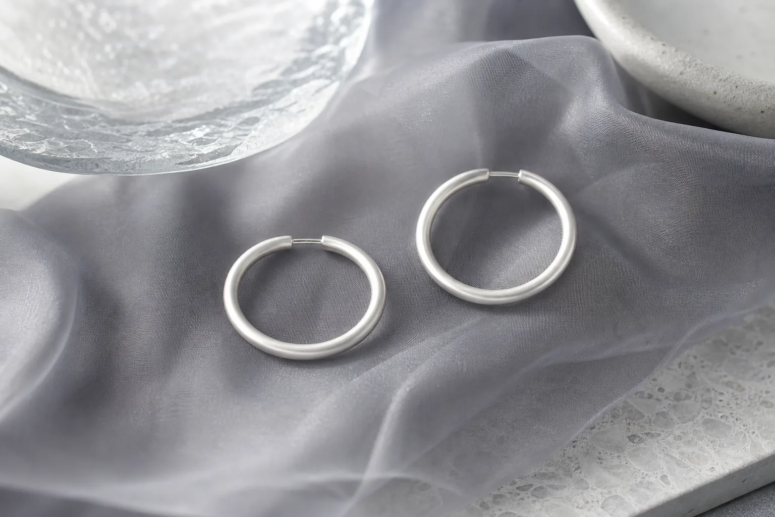 Pair of satin-frosted silver hoop earrings resting on smoky gray organza