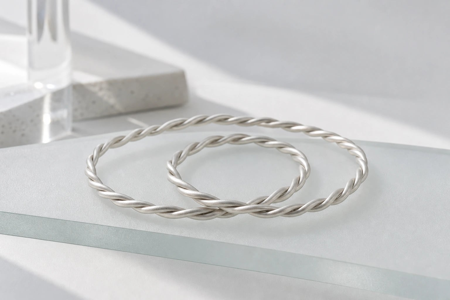 Two twisted silver bangles overlapping on a frosted glass ledge